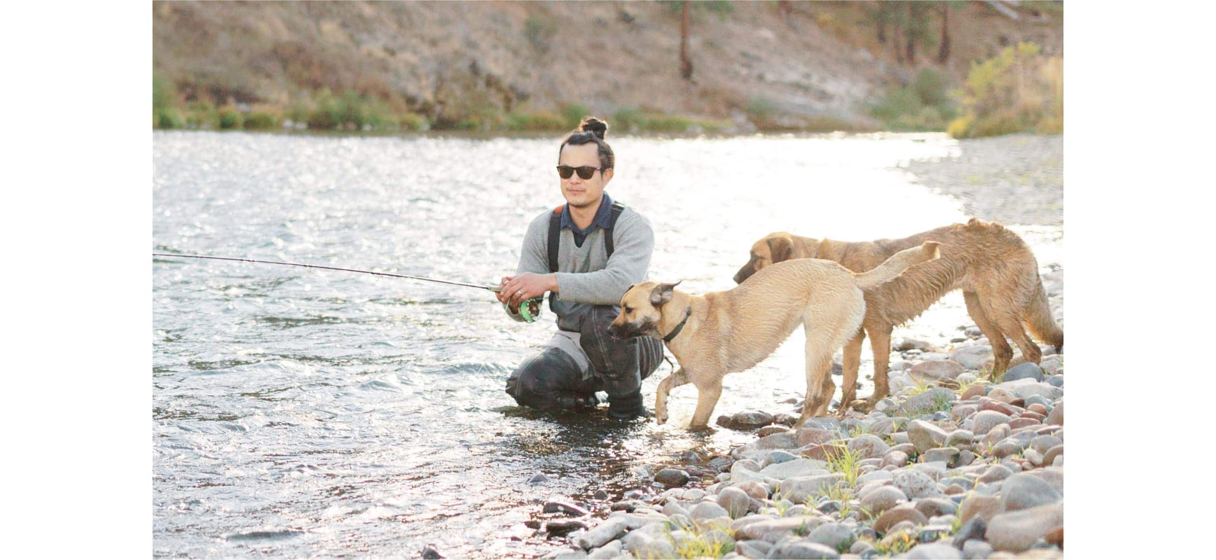  Burton crouches at the edge of a river fishing, as two dogs run around him. 